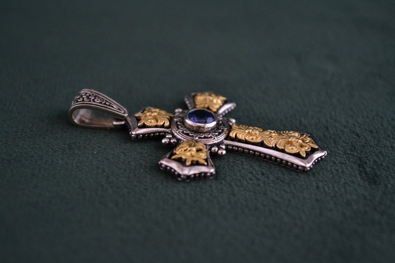 May include: A silver and gold cross pendant with a blue gemstone in the centre. The cross is decorated with intricate floral designs.