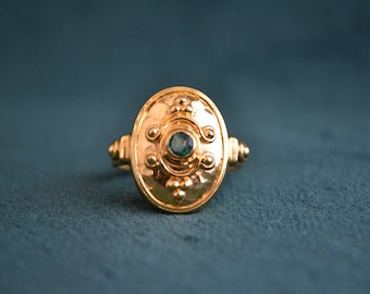 May include: A gold ring with a green gemstone set in a circular design. The ring has a textured surface and a raised edge.