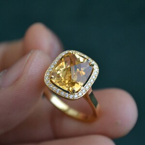 Pode incluir: Um anel de ouro com uma grande gema amarela de corte almofada, cercada por um halo de gemas menores e transparentes.