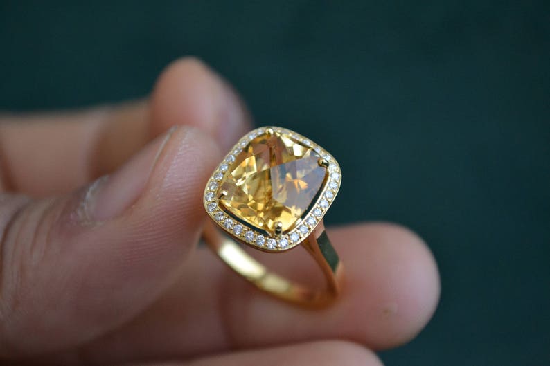 Pode incluir: Um anel de ouro com uma grande gema amarela de corte almofada, cercada por um halo de gemas menores e transparentes.