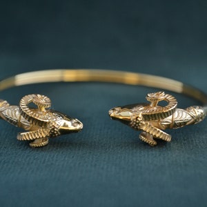 Puede incluir: Un brazalete de oro con detalles de cabeza de carnero. El brazalete está hecho de oro pulido y presenta detalles intrincados en las cabezas de carnero.