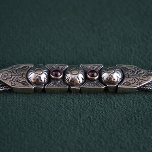 May include: A silver bracelet with a chain design and a decorative clasp featuring intricate floral patterns and red gemstones.