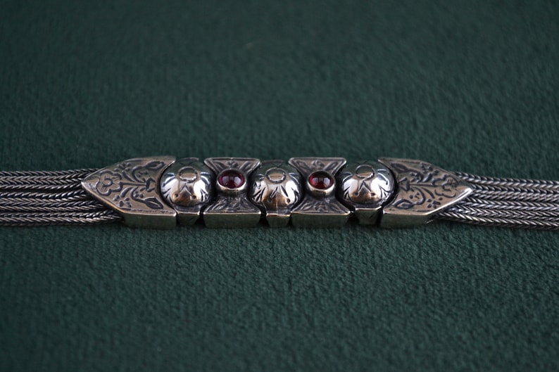 May include: A silver bracelet with a chain design and a decorative clasp featuring intricate floral patterns and red gemstones.
