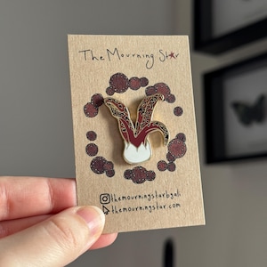 May include: A pin featuring a jester's hat design in red, gold, and white, set against a brown background with decorative brown circles. The text "The Mourning Star" is written at the top, with social media handles and a website address at the bottom.