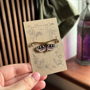 Puede incluir: Un broche de mariposa dorado con detalles en negro, blanco y burdeos, sujeto a una tarjeta decorativa. La tarjeta presenta el texto "The Mourning Star" e ilustraciones de hadas, flores y setas. El broche está sujeto en una mano.