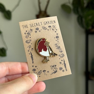 May include: Gold enamel pin featuring a red robin bird holding a key. The pin is on a brown card with the text "The Secret Garden" and "TMS" in black.