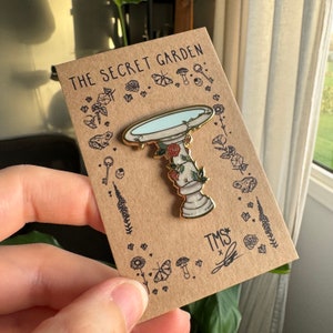 May include: Gold enamel pin depicting a white bird bath with a blue water basin and floral details. The pin is displayed on a brown card with the text "The Secret Garden" and "TMS*" in gold lettering.