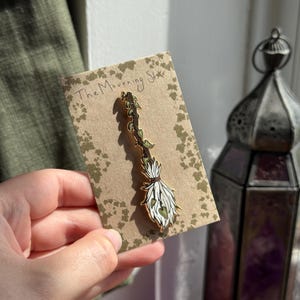 Puede incluir: Un pin dorado con forma de escoba con cerdas blancas y hojas verdes en forma de corazón. El pin está sujeto a una tarjeta marrón con el texto "The Mourning Star".