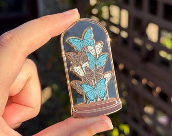 May include: A gold-toned enamel pin shaped like a glass cloche dome. Inside the dome are several butterflies in shades of bright blue, white, and brown. The base of the cloche is a solid brown colour, and the entire pin has a polished gold metal outline.