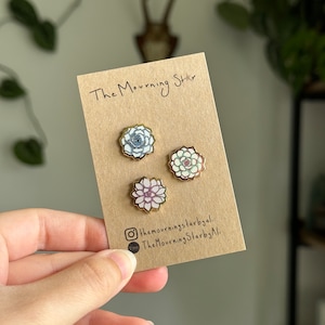 May include: Three gold succulent pins on a brown card with the text "The Mourning Star" and social media handles. The succulents are blue, white, and pink.