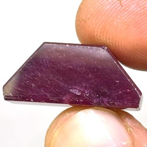 Könnte beinhalten: Ein tiefvioletter, facettierter Edelstein mit trapezförmiger Form. Der Edelstein hat eine glatte, polierte Oberfläche und wird zwischen zwei Fingern gehalten.