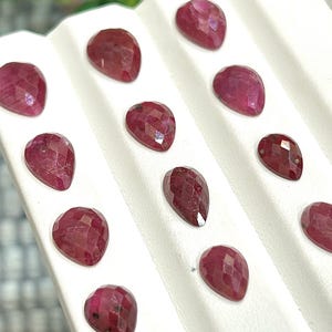 Puede incluir: Una colección de 12 piedras preciosas de rubí facetadas en forma de pera, expuestas en una bandeja blanca.