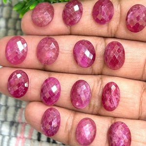 May include: A collection of 15 oval-shaped, faceted ruby gemstones in various shades of pink. The gemstones are arranged in a hand, showcasing their size and colour.