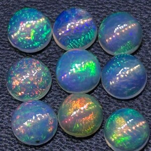 Peut inclure: Huit opales rondes taillées en cabochon disposées en grille. Chaque pierre précieuse présente une base translucide blanc laiteux avec des éclats vibrants de feu irisé vert, bleu et orange qui se déplacent sur les surfaces lisses et bombées.