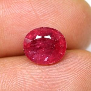 Op de afbeelding: Een dieprode ovale edelsteen met een facetgeslepen vorm. De edelsteen heeft een lichte glans en is geplaatst tegen een lichtroze achtergrond.