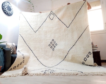 May include: A white Moroccan rug with black geometric designs. The rug has a fringe border and is being held up by two people.