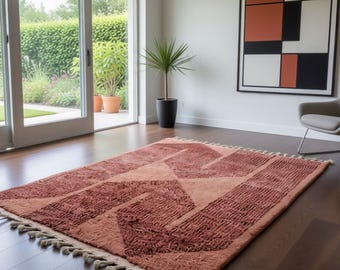Puede incluir: Una alfombra rectangular de color óxido con diseños geométricos en tonos melocotón y terracota. La alfombra, con borde de flecos, está colocada sobre un suelo de madera oscura. Una silla moderna y una obra de arte se ven al fondo.