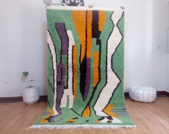 May include: A hand-woven wool rug with an abstract geometric design in shades of green, white, orange, black, and purple. The rug has a fringed border.