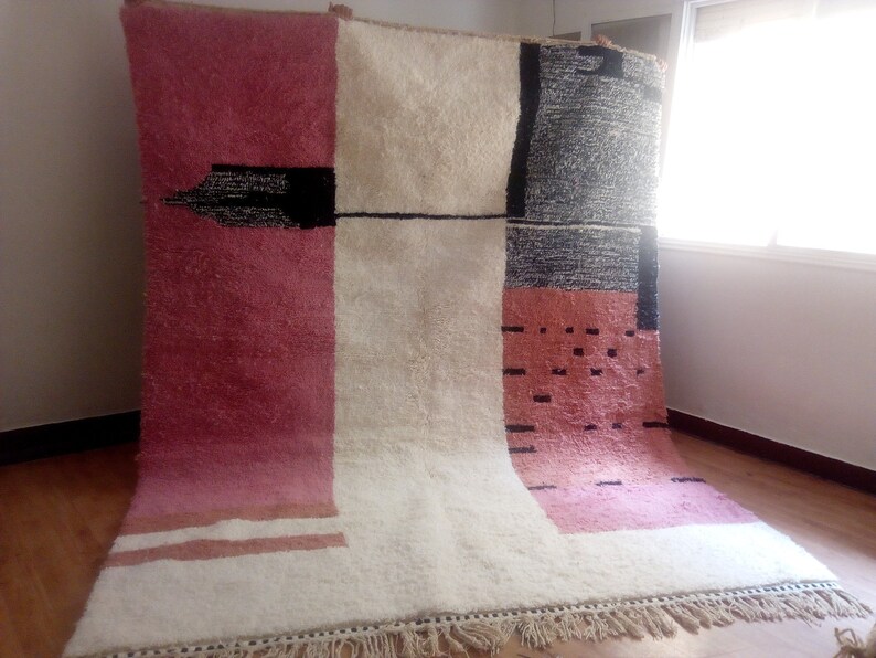 May include: A large, rectangular rug with a geometric pattern in shades of pink, white, and black. The rug has a fringe border.