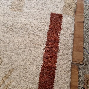 May include: Close-up of a textured rug with a cream-colored background and a bold, rust-colored stripe. The rug's design includes abstract shapes in shades of beige and brown. The rug is made of a soft, plush material, perfect for adding warmth and style to any room.