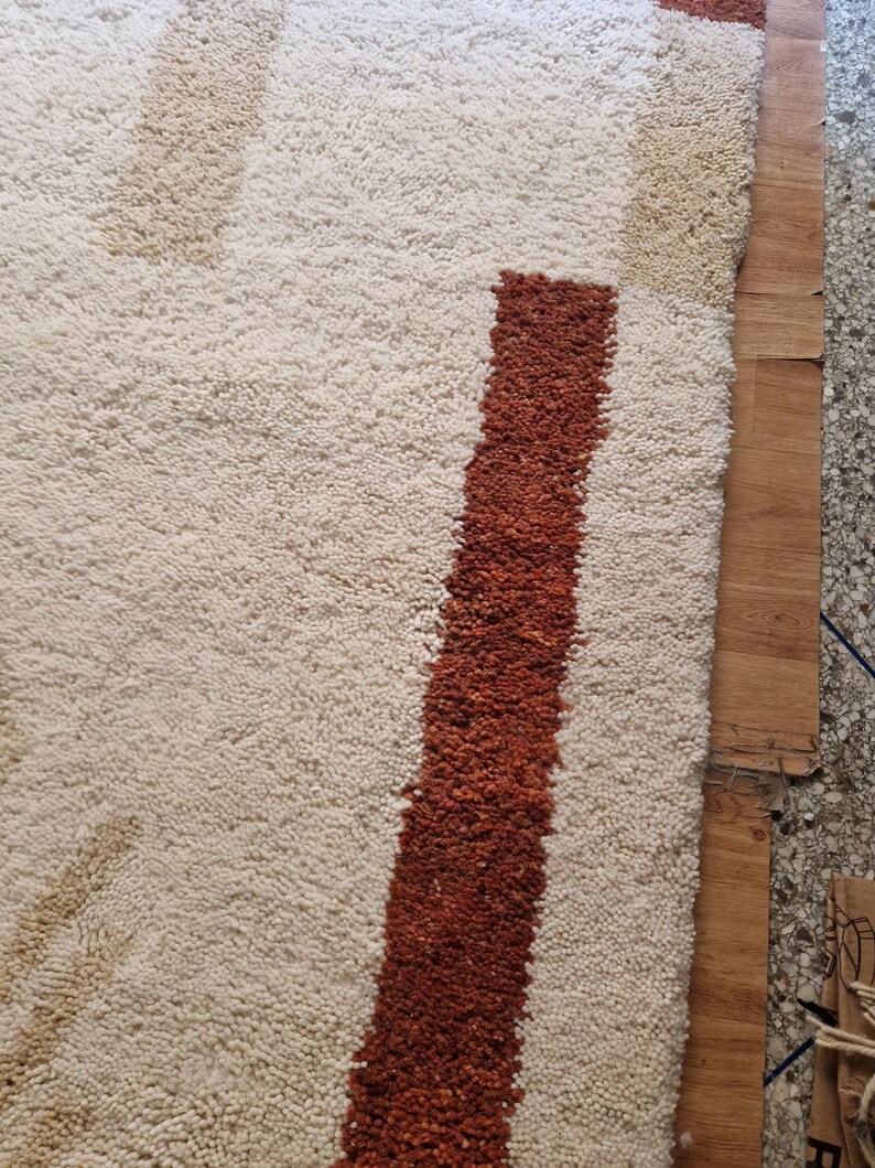 May include: Close-up of a textured rug with a cream-colored background and a bold, rust-colored stripe. The rug's design includes abstract shapes in shades of beige and brown. The rug is made of a soft, plush material, perfect for adding warmth and style to any room.