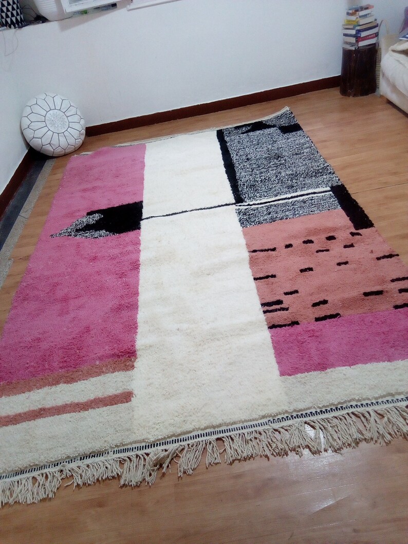 May include: A white, pink, and black geometric patterned rug with fringe. The rug has a modern design with abstract shapes and lines.