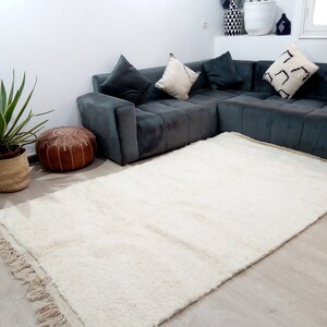 Pode incluir: Um tapete branco de pelo alto com franjas nas bordas, colocado em uma sala de estar com um sof&aacute; seccional cinza e um pufe de couro marrom.