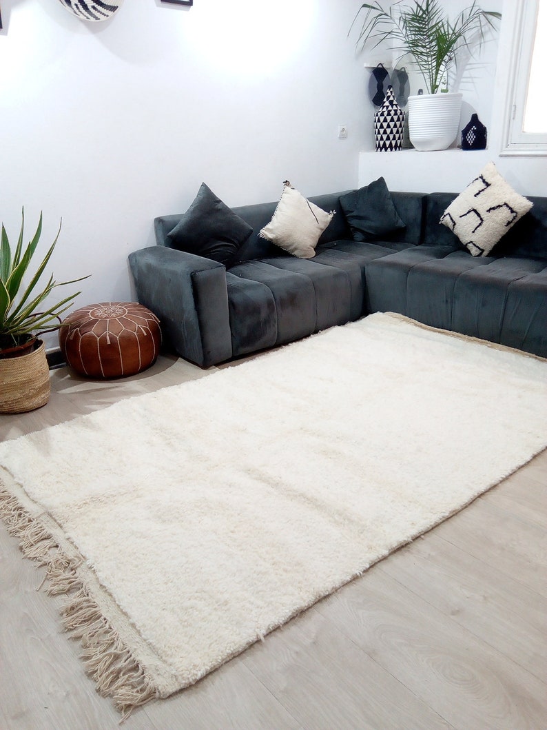 Pode incluir: Um tapete branco de pelo alto com franjas nas bordas, colocado em uma sala de estar com um sof&aacute; seccional cinza e um pufe de couro marrom.