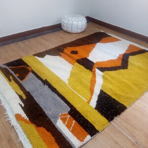 May include: A rectangular rug with a geometric design in shades of yellow, orange, brown, gray, and white. The rug has a textured surface and a fringe along one edge. A white leather pouf is in the background.