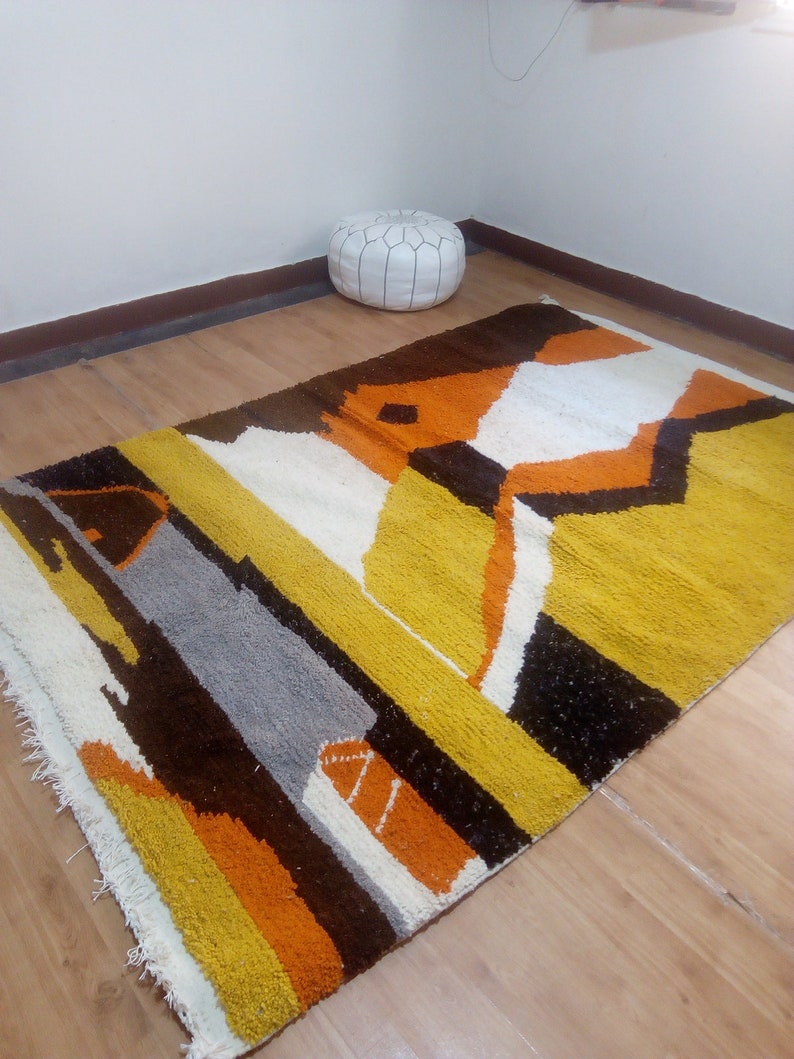 May include: A rectangular rug with a geometric design in shades of yellow, orange, brown, gray, and white. The rug has a textured surface and a fringe along one edge. A white leather pouf is in the background.