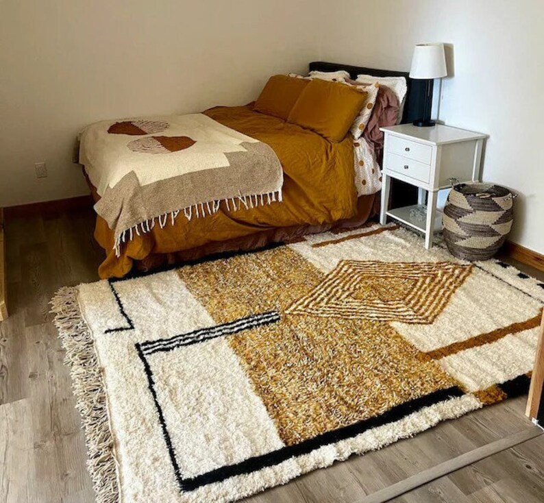 Puede incluir: Una alfombra blanca y marr&oacute;n con un dise&ntilde;o geom&eacute;trico en tonos marrones y beige. La alfombra se coloca delante de una cama con una manta y almohadas de estampado marr&oacute;n y beige.