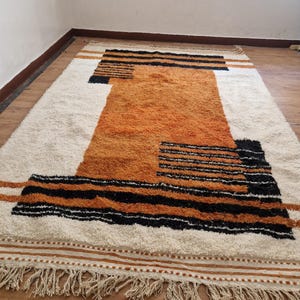 May include: A white and orange geometric patterned rug with black stripes and fringe. The rug has a modern design and is made of a soft, fluffy material.
