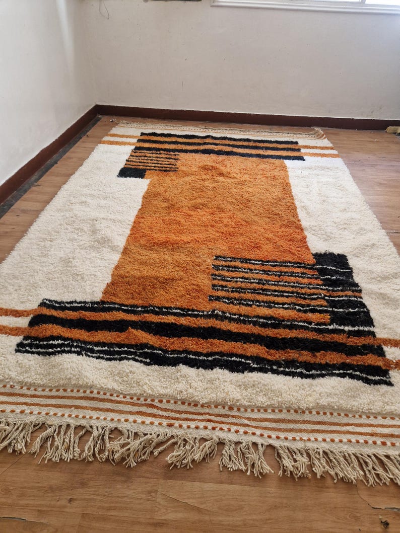 May include: A white and orange geometric patterned rug with black stripes and fringe. The rug has a modern design and is made of a soft, fluffy material.
