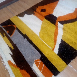 May include: A rectangular rug with an abstract geometric design. The rug features a combination of colors, including yellow, orange, white, gray, and brown. The rug is placed on a wooden floor.