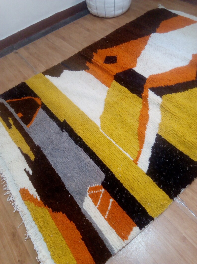 May include: A rectangular rug with an abstract geometric design. The rug features a combination of colors, including yellow, orange, white, gray, and brown. The rug is placed on a wooden floor.