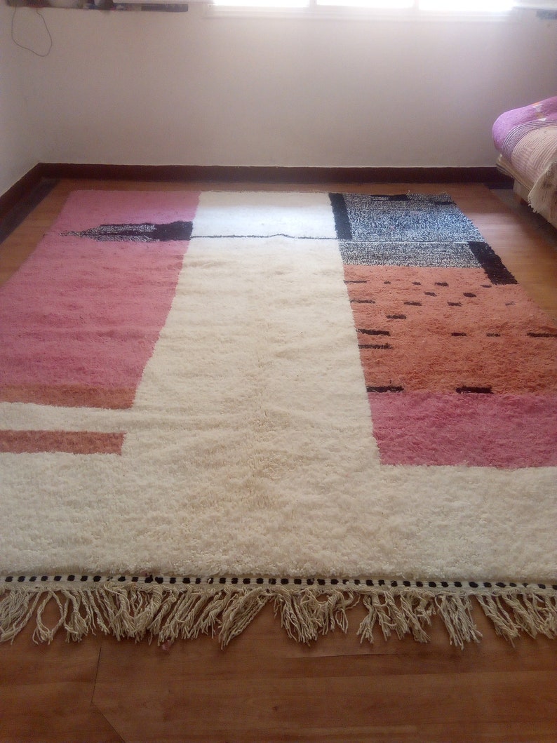 May include: A white, pink, and orange geometric patterned rug with a black fringe border. The rug has a white center with pink and orange sections on either side.