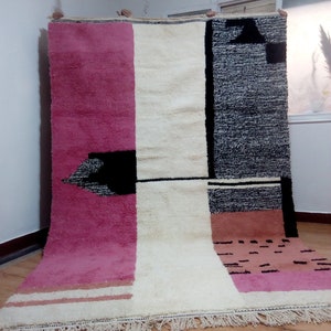 May include: A large, rectangular rug with a geometric design. The rug is made of wool and has a fringe border. The rug is in shades of pink, white, black, and brown.