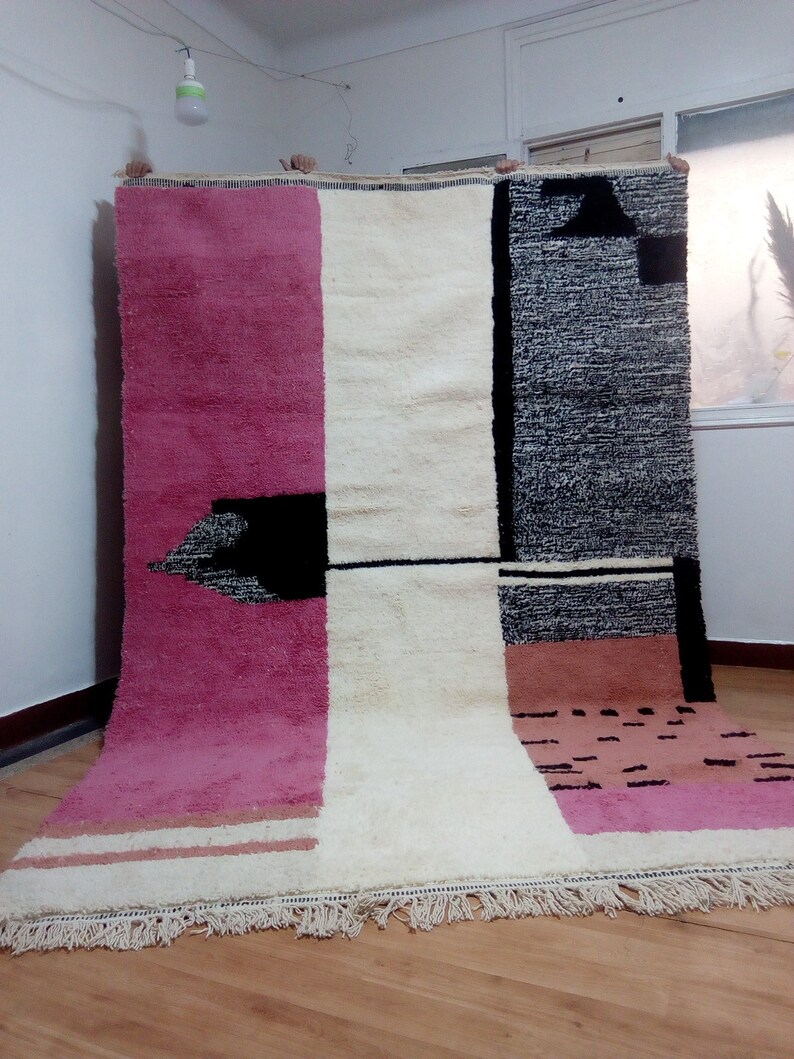 May include: A large, rectangular rug with a geometric design. The rug is made of wool and has a fringe border. The rug is in shades of pink, white, black, and brown.