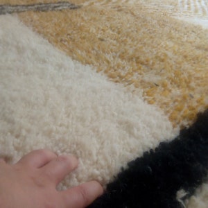 Puede incluir: Primer plano de una alfombra de pelo blanco, amarillo y negro. La alfombra tiene un patr&oacute;n geom&eacute;trico y est&aacute; hecha de fibras suaves y esponjosas.