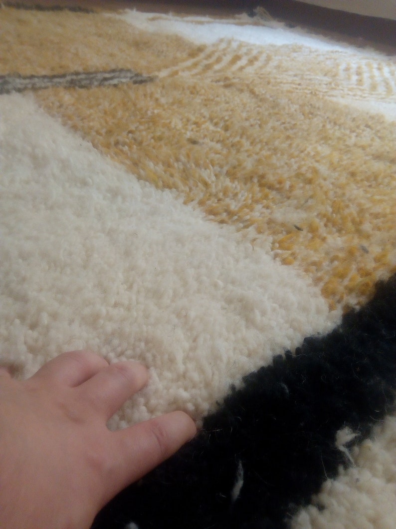 Puede incluir: Primer plano de una alfombra de pelo blanco, amarillo y negro. La alfombra tiene un patr&oacute;n geom&eacute;trico y est&aacute; hecha de fibras suaves y esponjosas.