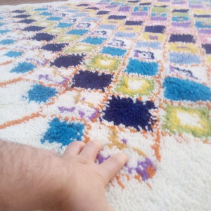 May include: A white rug with a colorful geometric pattern. The rug is made of fluffy, soft fibers and features squares of blue, green, yellow, orange, and purple.
