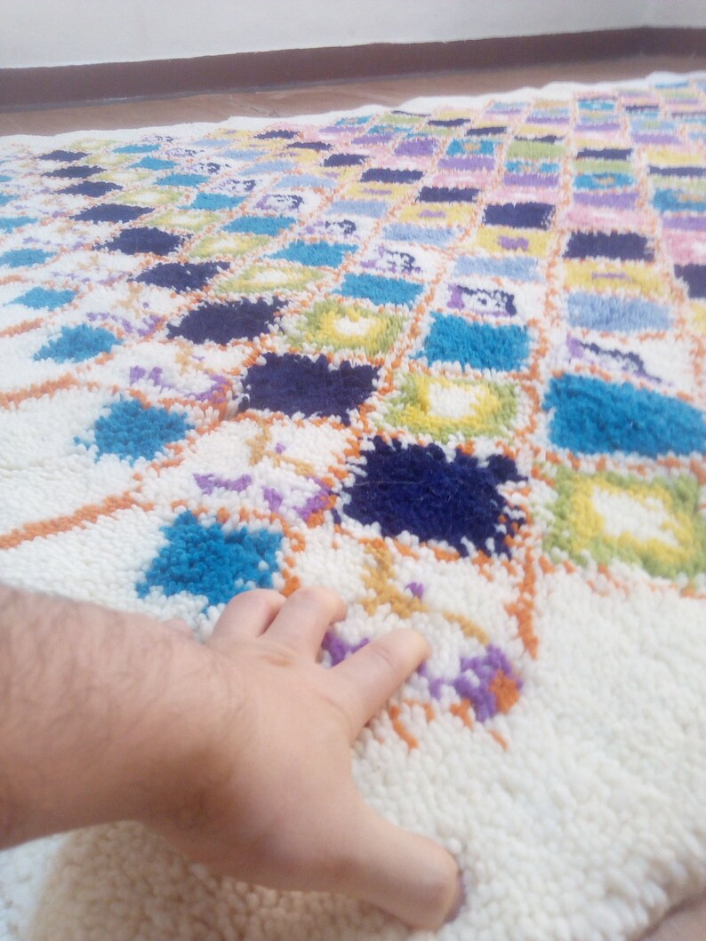 May include: A white rug with a colorful geometric pattern. The rug is made of fluffy, soft fibers and features squares of blue, green, yellow, orange, and purple.