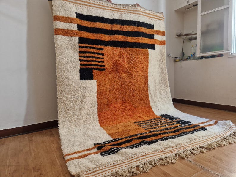 May include: A white, orange, and black geometric patterned rug with a fringe border. The rug has a modern design and is made of a soft, fluffy material.