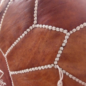 May include: A brown leather pouf with white braided trim and tassels. The pouf is made of multiple leather panels stitched together.