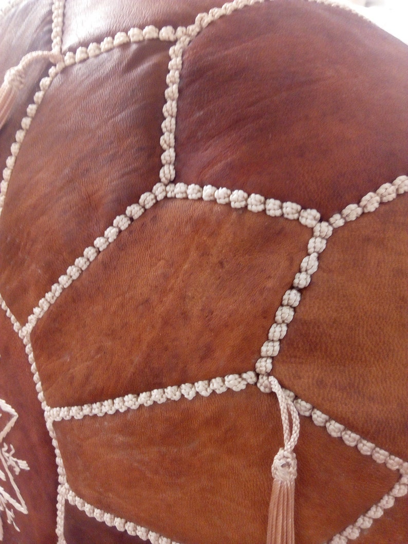 May include: A brown leather pouf with white braided trim and tassels. The pouf is made of multiple leather panels stitched together.