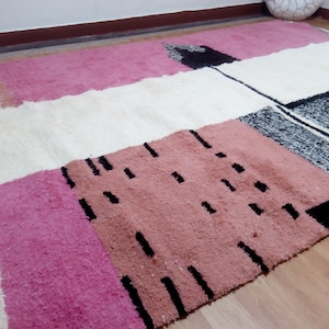 May include: A white, pink, and black geometric patterned rug with a fluffy texture. The rug has a modern design with abstract shapes and lines.