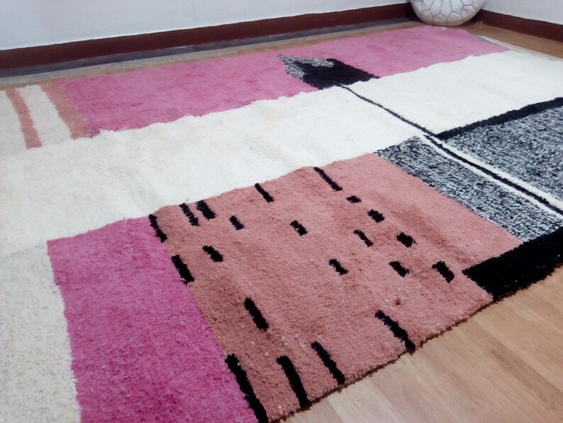 May include: A white, pink, and black geometric patterned rug with a fluffy texture. The rug has a modern design with abstract shapes and lines.