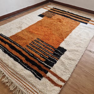 May include: A white and orange geometric patterned rug with black accents and fringe on the edges.
