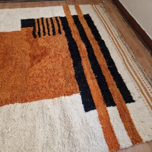 May include: A white, orange, and black geometric patterned rug with a shaggy texture. The rug has a white background with orange and black stripes and geometric shapes.