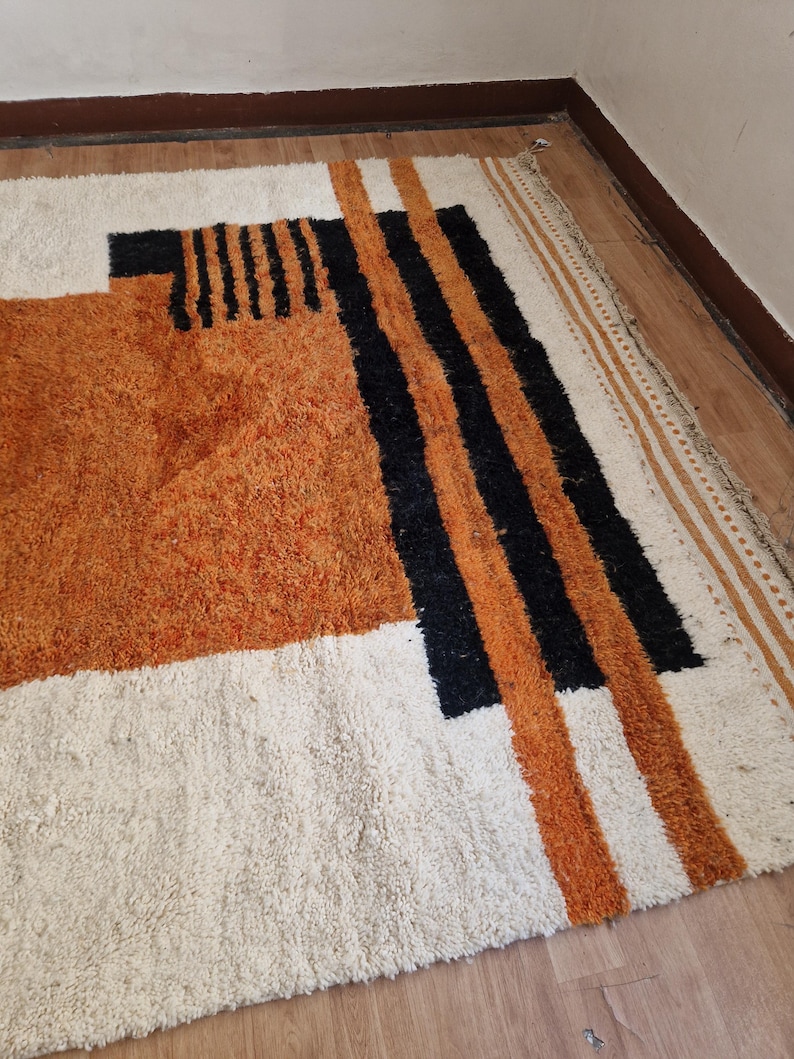 May include: A white, orange, and black geometric patterned rug with a shaggy texture. The rug has a white background with orange and black stripes and geometric shapes.